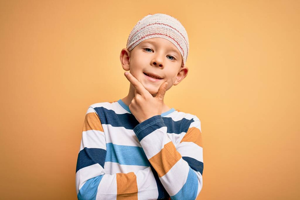 Маленький белый мальчик, раненый в медицинскую повязку на голове на жёлтом фоне, уверенно смотрит на камеру, улыбаясь со скрещенными руками и поднятой на подбородок рукой. Мысли позитивно
. - Фото, изображение