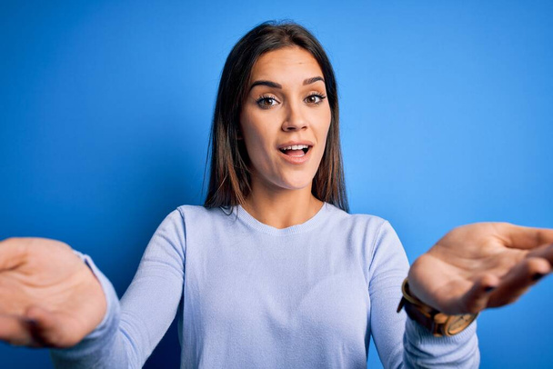 Jonge mooie brunette vrouw dragen casual trui staan over blauwe achtergrond glimlachend vrolijk aanbieden van handen geven hulp en acceptatie. - Foto, afbeelding