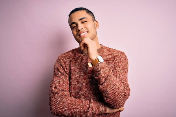 Young brazilian man wearing casual sweater standing over isolated pink background looking confident at the camera with smile with crossed arms and hand raised on chin. Thinking positive. - Фото, изображение