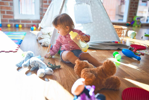 Очаровательный малыш держит бутылочку для кормления вокруг кучи игрушек в детском саду
 - Фото, изображение