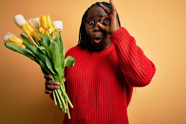 Mladá Afroameričanka plus velká žena s copánky držícími kytici žlutých tulipánů, která dělá v pořádku gesto šokované překvapeným obličejem, oči hledící skrz prsty. Nevěřící výraz. - Fotografie, Obrázek