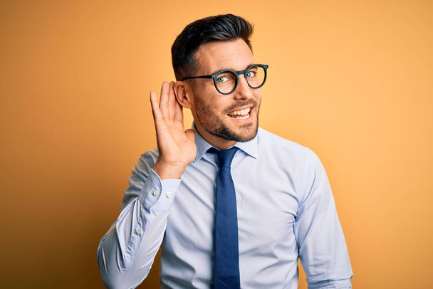 Young handsome businessman wearing tie and glasses standing over yellow background smiling with hand over ear listening an hearing to rumor or gossip. Deafness concept. - Photo, Image