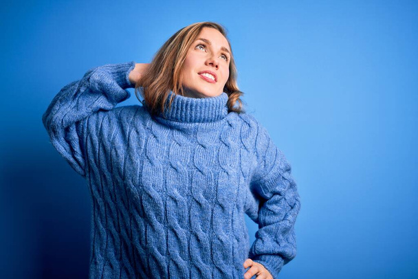 Genç, sarışın bir kadın mavi arka plan üzerine balıkçı yaka kazak giyiyor yorgun ve rahat, uykulu ve sabahın erken saatleri için esniyor. - Fotoğraf, Görsel