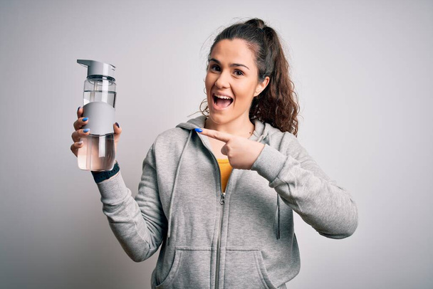 Beautiful woman with curly hair doing sport drinking bottle of water over white background very happy pointing with hand and finger - Фото, зображення