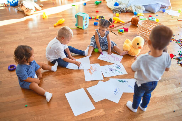 Очаровательная группа малышей, сидящих на полу и рисующих бумагой и карандашом вокруг большого количества игрушек в детском саду
 - Фото, изображение