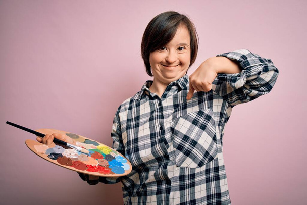 Junge Frau mit Down-Syndrom hält Malerpalette und Pinsel über rosa Hintergrund mit Überraschungsgesicht und zeigt mit dem Finger auf sich selbst - Foto, Bild
