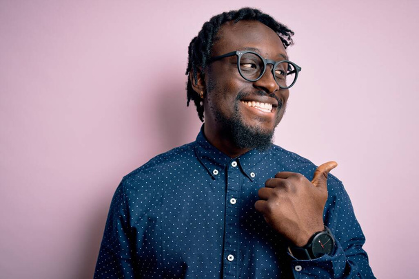 Junger gutaussehender afrikanisch-amerikanischer Mann mit lässigem Hemd und Brille vor rosa Hintergrund, lächelnd mit glücklichem Gesicht, das mit erhobenem Daumen zur Seite zeigt. - Foto, Bild
