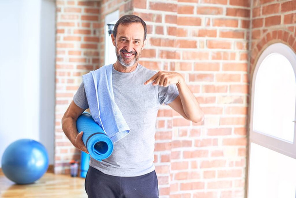 Красивий спортсмен середнього віку тримає килимок і рушник стоячи перед тренуванням у спортзалі з несподіваним обличчям, вказуючи пальцем на себе
 - Фото, зображення