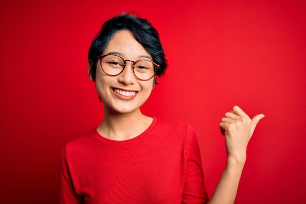 Молода красива азіатська дівчина в повсякденній футболці і окулярах над ізольованим червоним тлом посміхається з щасливим обличчям дивлячись і вказуючи на бік з великим пальцем вгору
. - Фото, зображення