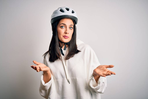 Mavi gözlü genç bisikletçi kadın izole edilmiş beyaz arka plan üzerinde bisiklet kaskı takıyor ve elleri ve kolları havada kafası karışmış bir ifade takınıyor. Şüphe kavramı. - Fotoğraf, Görsel