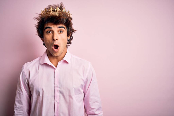 Młody przystojny mężczyzna ubrany w królewską koronę stojący nad odizolowanym różowym tle przerażony i zszokowany zaskakującą ekspresją, strachem i podnieconą twarzą. - Zdjęcie, obraz