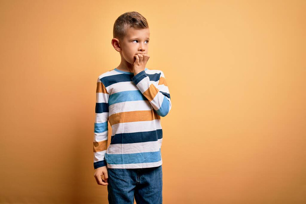 Jeune enfant caucasien aux yeux bleus portant une chemise rayée colorée sur fond jaune, stressé et nerveux avec les mains sur les ongles mordants de la bouche. Problème d'anxiété
. - Photo, image