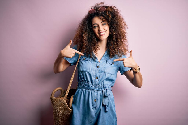 Young beautiful woman with curly hair and piercing wearing denim dress and wicker bag looking confident with smile on face, pointing oneself with fingers proud and happy. - Photo, Image