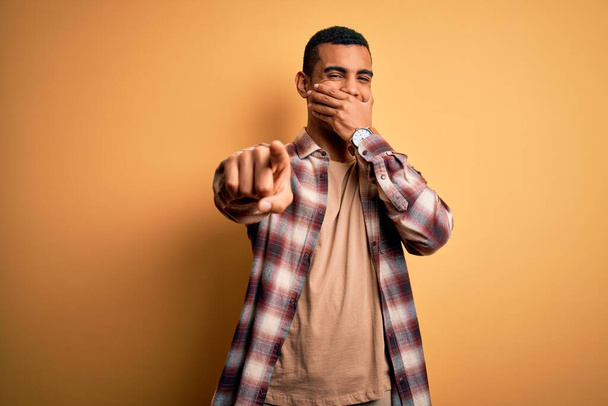 Joven hombre afroamericano guapo usando camisa casual de pie sobre fondo amarillo riéndose de ti, señalando con el dedo a la cámara con la mano sobre la boca, expresión de vergüenza
 - Foto, imagen