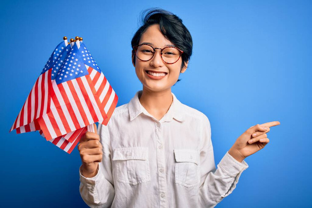 Молода красива азіатська патріотична дівчина тримає об'єднані держави прапори святкування Дня незалежності дуже щаслива вказуючи рукою і пальцем на бік
 - Фото, зображення