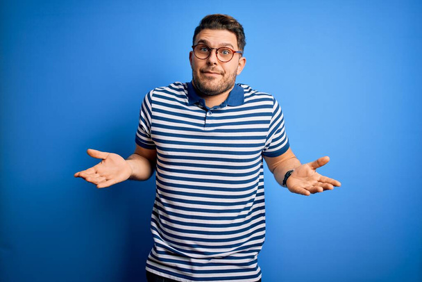 Young man with blue eyes wearing glasses and casual striped t-shirt over blue background clueless and confused expression with arms and hands raised. Doubt concept. - Photo, Image