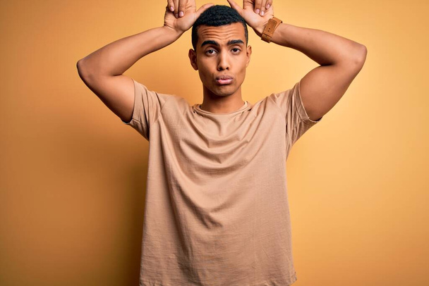 Young handsome african american man wearing casual t-shirt standing over yellow background doing funny gesture with finger over head as bull horns - Foto, afbeelding
