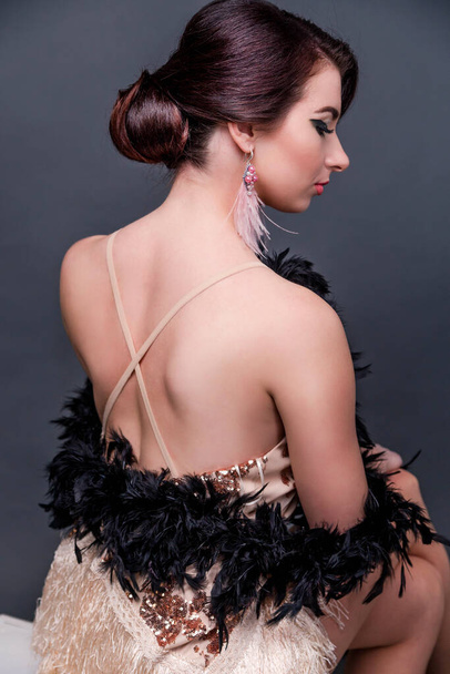 Beautiful brunette  woman in boa from ostrich feathers sits in front of the camera with his back and posing. Vertycal photo - Фото, изображение