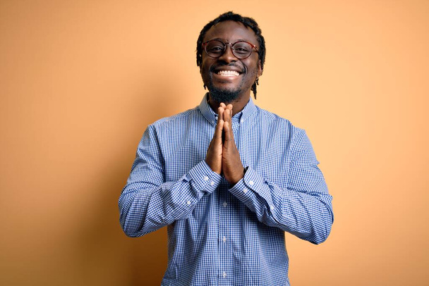 Young handsome african american man wearing shirt and glasses over yellow background praying with hands together asking for forgiveness smiling confident. - Photo, Image