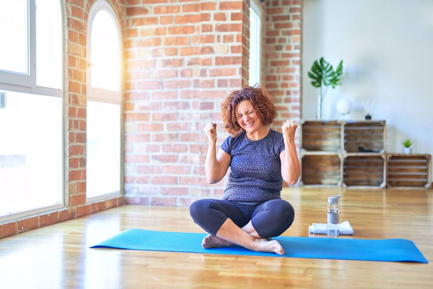 Middle age beautiful sportswoman wearing sportswear sitting on mat practicing yoga at home very happy and excited doing winner gesture with arms raised, smiling and screaming for success. Celebration concept. - Photo, Image