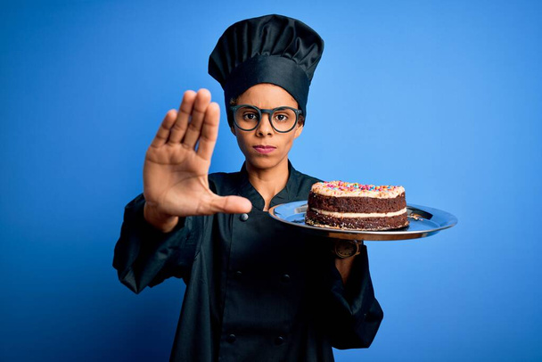 Joven panadera afroamericana con uniforme de cocina y sombrero sosteniendo pastel con la mano abierta haciendo stop sign con expresión seria y confiada, gesto de defensa
 - Foto, Imagen