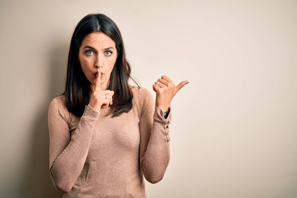 Young brunette woman with blue eyes wearing casual sweater over isolated white background asking to be quiet with finger on lips pointing with hand to the side. Silence and secret concept. - Foto, Imagem