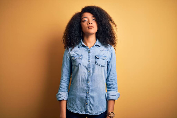 Νεαρή όμορφη Αφροαμερικανή γυναίκα με αφρο μαλλιά που στέκεται πάνω από κίτρινο απομονωμένο φόντο Χαλαρωμένη με σοβαρή έκφραση στο πρόσωπο. Απλή και φυσική κοιτάζοντας την κάμερα. - Φωτογραφία, εικόνα