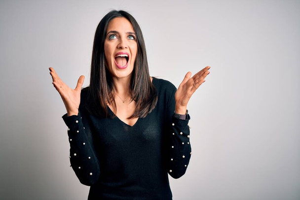 Young brunette woman with blue eyes wearing black casual sweater over white background celebrating mad and crazy for success with arms raised and closed eyes screaming excited. Winner concept - Fotoğraf, Görsel