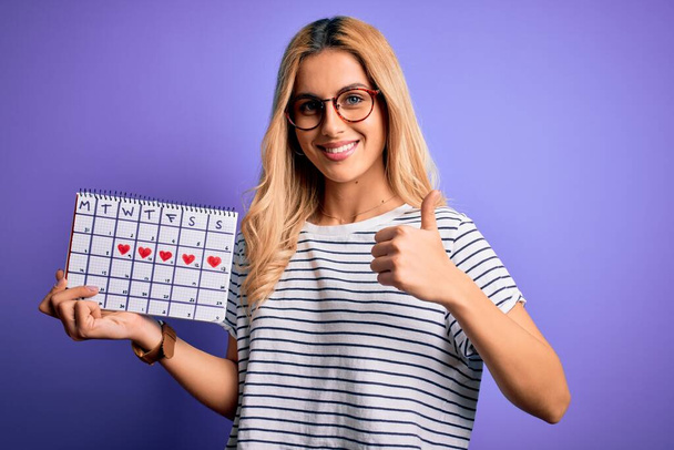 Young beautiful blonde woman holding period calendar to control menstrual cycle happy with big smile doing ok sign, thumb up with fingers, excellent sign - Fotografie, Obrázek