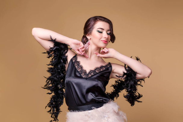 Brunette beautiful woman in black boa from ostrich feathers is dancing on beige background. Horizontal photo - Фото, изображение