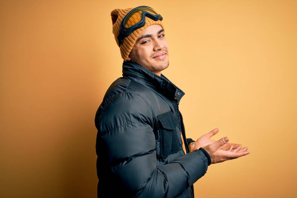 雪のスポーツウェアとスキーゴーグルを身に着けている若いブラジルのスキー男は黄色の背景の上に脇を指している手を開いてヤシの木は、コピースペースを示す広告笑顔興奮幸せな興奮を提示 - 写真・画像