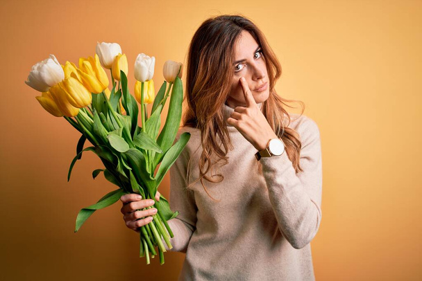 Young beautiful brunette woman holding bouquet of yellow tulips over isolated background Pointing to the eye watching you gesture, suspicious expression - Photo, Image