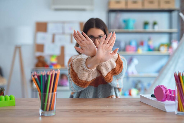 Молодая красивая учительница в свитере и очках сидит на столе в детском саду Отказ выражение лица скрещивание рук и ладоней делает негативный знак, сердитый лицо
 - Фото, изображение