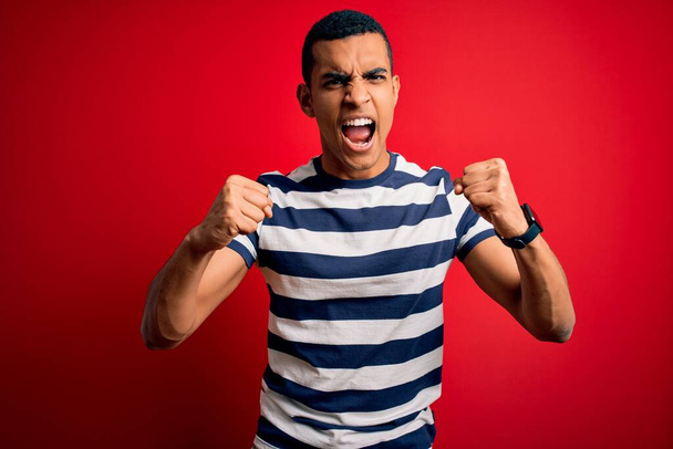 Handsome african american man wearing casual striped t-shirt standing over red background angry and mad raising fists frustrated and furious while shouting with anger. Rage and aggressive concept. - Foto, imagen