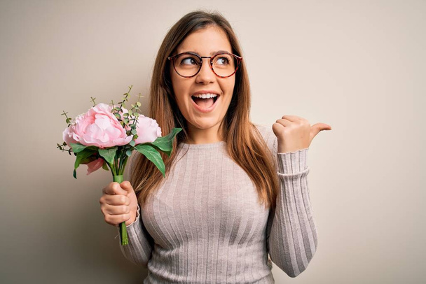 Young blonde woman holding beautiful romantic floral bouquet over isolated background pointing and showing with thumb up to the side with happy face smiling - Photo, Image