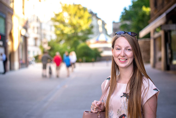 Young cheerful woman with long hair and freckles smiling at camera while having a walk through the streets of an european town. Summer season. Elegant woman, wearing a purse.  - Foto, Bild