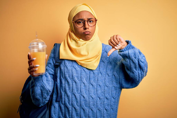 Молода афро-американська туристка в муслім хіджаб п'є апельсиновий сік з сердитим обличчям, негативний знак, що показує неприязнь великим пальцями вниз, відторгнення концепції - Фото, зображення