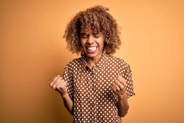 Junge schöne afrikanisch-amerikanische Frau in lässigem Hemd vor gelbem Hintergrund sehr glücklich und aufgeregt dabei Siegergeste mit erhobenen Armen, lächelnd und schreiend nach Erfolg. Festkonzept. - Foto, Bild