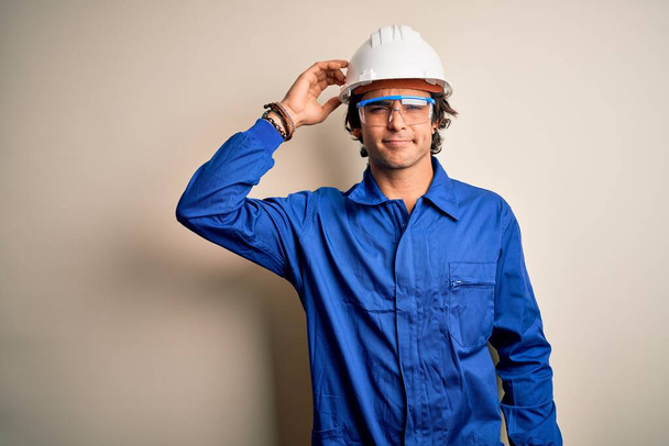 Young constructor man wearing uniform and security helmet over isolated white background confuse and wonder about question. Uncertain with doubt, thinking with hand on head. Pensive concept. - Foto, imagen
