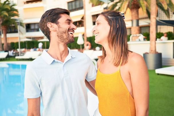 Молодая красивая пара в отпуске улыбается счастливой и уверенной. Стоя с улыбкой на лице обнимаясь в пляжном клубе
 - Фото, изображение
