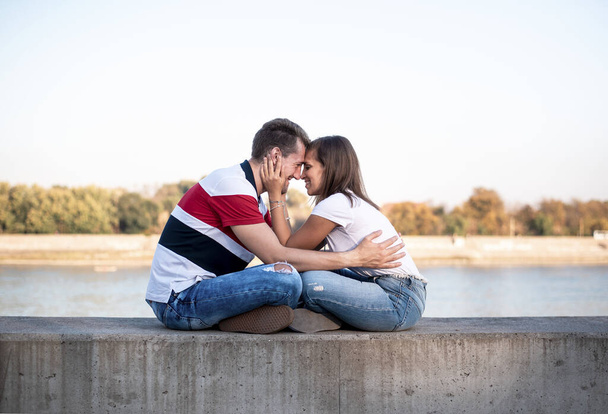 Vista lateral de una feliz pareja amorosa joven y casual en jeans, sentados uno frente al otro y sujetándose suavemente en un muelle del río
. - Foto, imagen