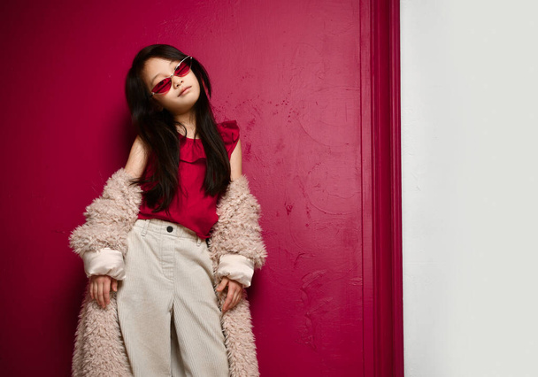Cool asian kid girl in narrow sunglasses and wide pants is standing in slipping off her shoulders beige fur coat - Foto, immagini