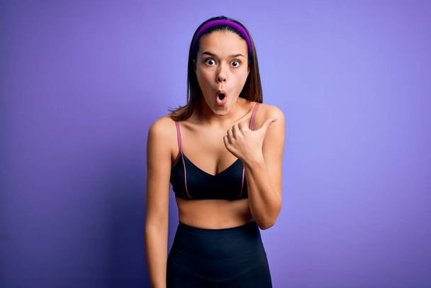 Young beautiful sporty girl doing sport wearing sportswear over isolated purple background Surprised pointing with hand finger to the side, open mouth amazed expression. - Photo, Image