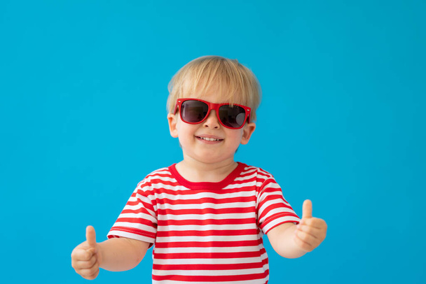 Lähikuva muotokuva onnellinen lapsi yllään raidallinen paita sinistä taustaa vasten. Lapsella on hauskaa kesälomalla. Poika näyttää peukalonsa - Valokuva, kuva
