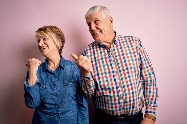 Älteres schönes Paar, das zusammen vor isoliertem rosa Hintergrund steht, lächelnd mit glücklichem Gesicht schaut und mit erhobenem Daumen zur Seite zeigt. - Foto, Bild