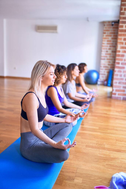 Junge schöne Gruppe von Sportlerinnen, die glücklich lächelnd Yoga praktizieren. Sitzen bei Lotus-Pose im Fitnessstudio - Foto, Bild