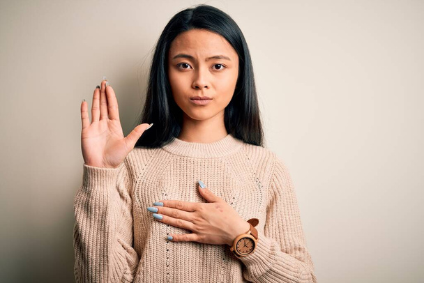 Молодая красивая китаянка в обычном свитере на изолированном белом фоне, ругаясь рукой на груди и открытой ладони, дает клятву верности
 - Фото, изображение