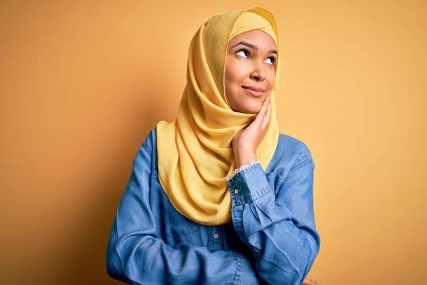 巻き毛を持つ若い美しい女性は、質問について考えて顎の手で黄色の背景にアラブの伝統的なヒジャーブを身に着けています,集中的な表現.笑顔と思慮深い顔。疑いの概念. - 写真・画像