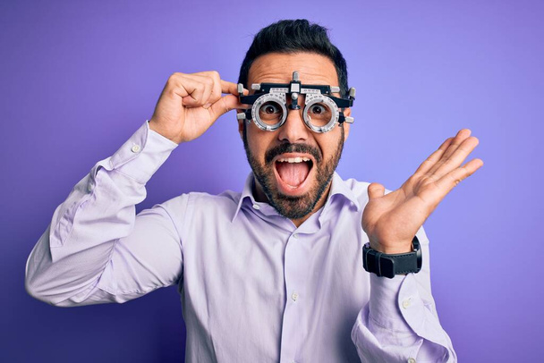 Young handsome optical man with beard wearing optometry glasses over purple background very happy and excited, winner expression celebrating victory screaming with big smile and raised hands - Foto, afbeelding
