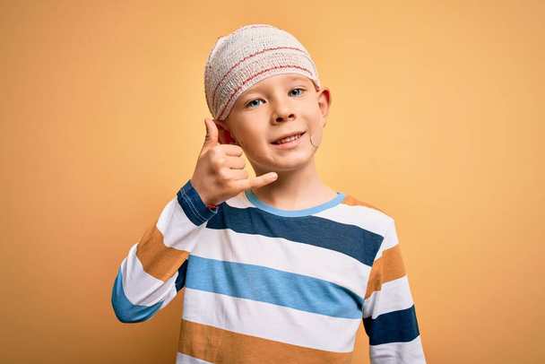 Young little caucasian kid injured wearing medical bandage on head over yellow background smiling doing phone gesture with hand and fingers like talking on the telephone. Communicating concepts. - Foto, imagen